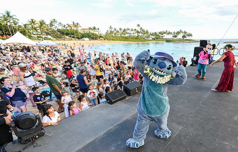 Lilo on stage with Mickey Mouse and audience of children with beach and lagoon in the background.