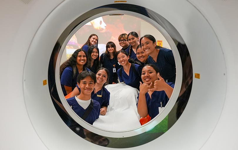 High school students in scrubs smiling through circular opening of a CT scanner.