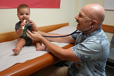 Smiling pediatric patient sitting on an exam table, holding a tongue depressor with doctor listening to his lungs.